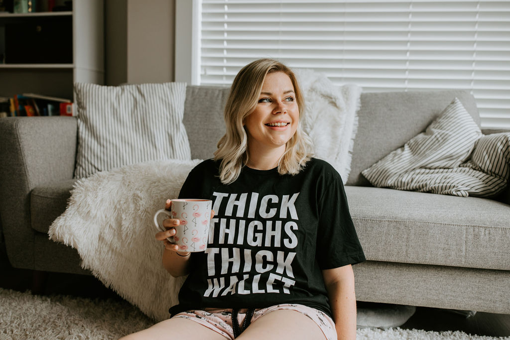 Body Image context, Melissa Rodgers sitting on living room floor in t-shirt that reads Thick Thighs Thick Wallet
