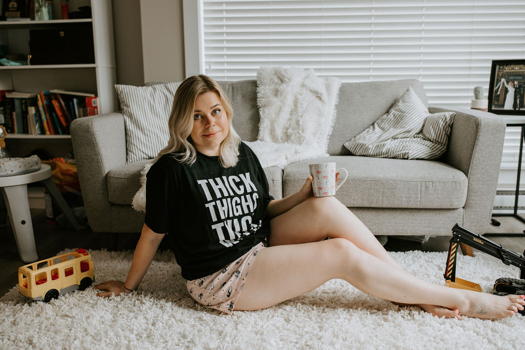 Body Image context, Melissa Rodgers sits on floor in t-shirt that reads Thick Thighs Thick Wallet