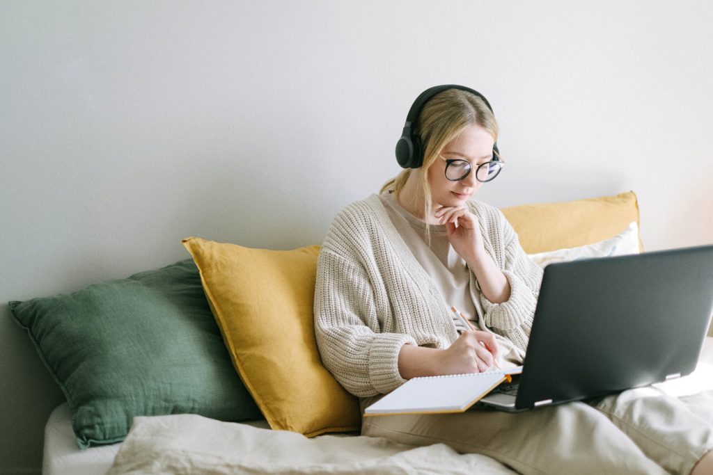Online Course context, woman sitting in bed on laptop with headphones and writing in notebook